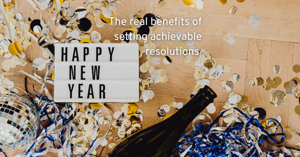 the remnants of a new years eve party with gold confetti an empty chapage bottle and a light box with happy new year and text which says the real benefits of setting achievable resolutions